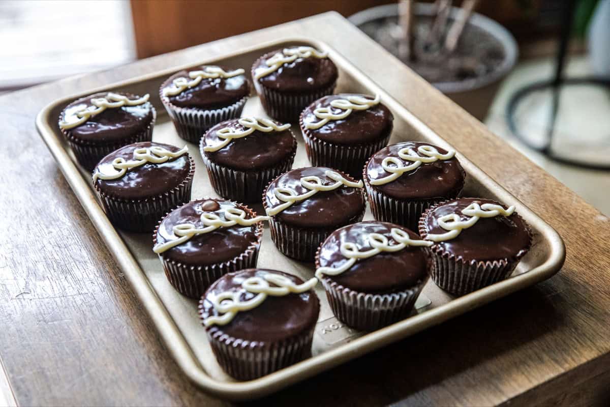 A baking tray with twelve chocolate cupcakes, each topped with a swirl of white icing, sits on a wooden surface indoors.