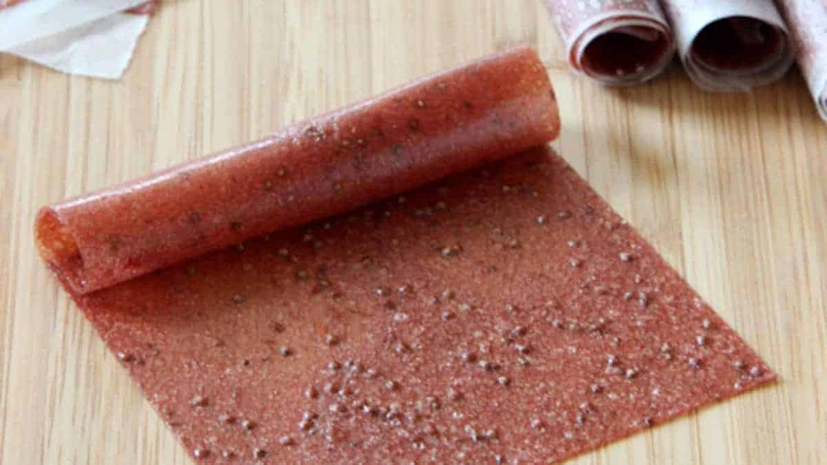 A sheet of homemade fruit leather is being rolled up on a wooden surface, with additional rolled fruit leather pieces in the background.