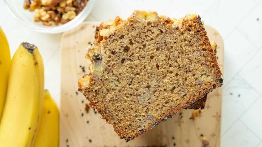 A sliced loaf of banana bread with walnuts on top, placed on a wooden cutting board next to bananas and a bowl of walnuts.