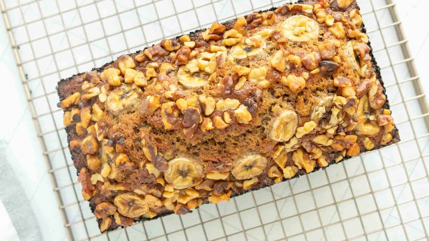 A loaf of banana bread topped with banana slices and chopped walnuts, cooling on a wire rack.