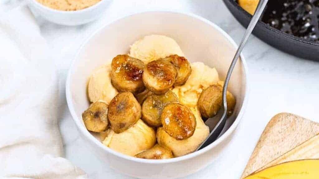 A bowl of vanilla ice cream topped with caramelized banana slices, with a spoon in the bowl. A partial view of a cutting board and a skillet are in the background.