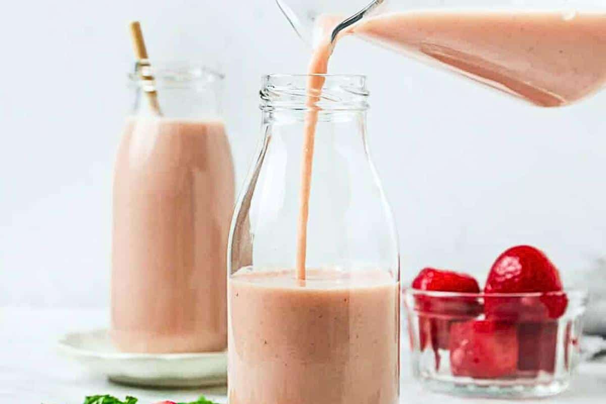 A pink smoothie is being poured into a clear glass bottle, with another filled bottle and a bowl of strawberries in the background.