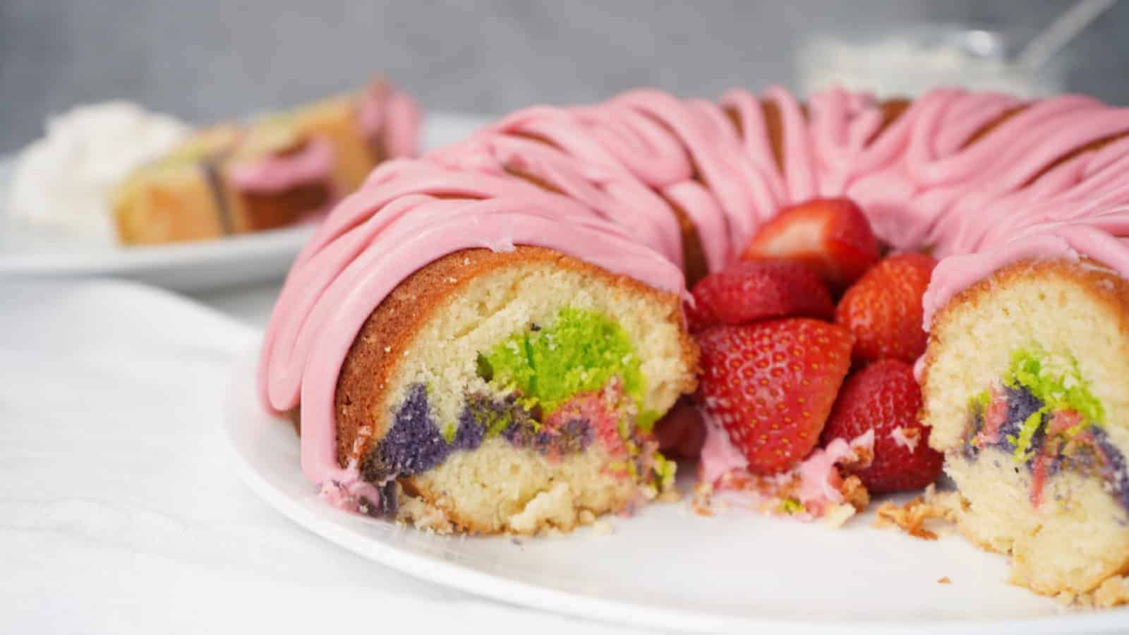 A bundt cake with pink icing, filled with strawberries and sections of green and purple cake, sits on a white plate with a slice served in the background.