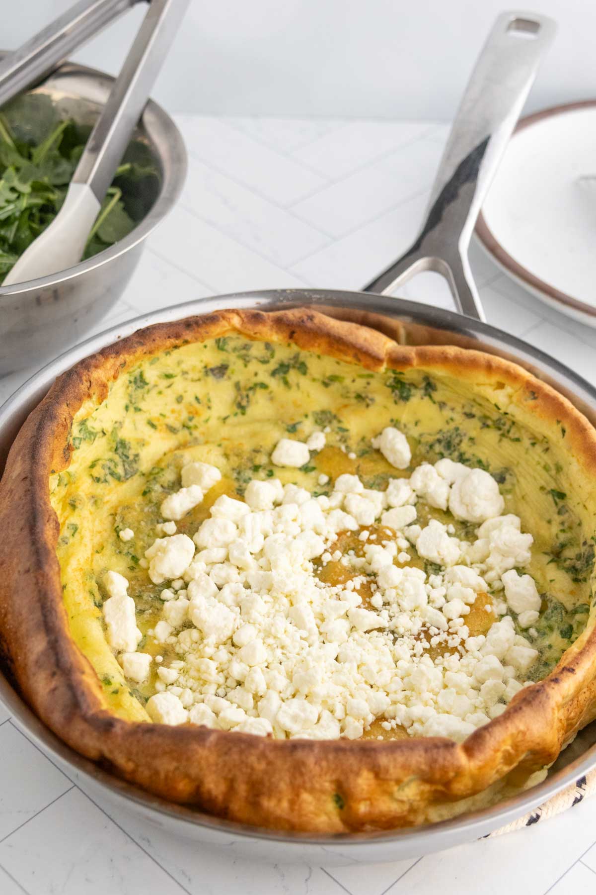 A baked Dutch baby pancake with herbs and crumbled cheese on top sits in a skillet next to a bowl of greens and a plate.