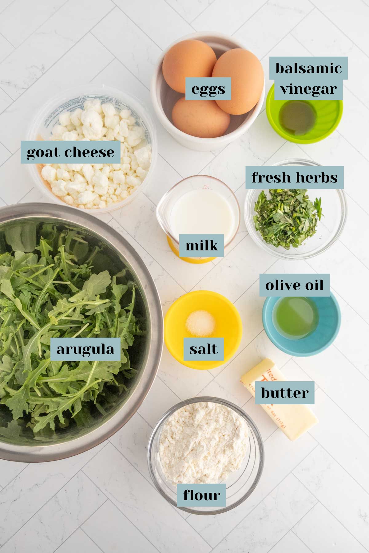Top-down view of ingredients in bowls for a recipe, including eggs, arugula, goat cheese, flour, milk, butter, fresh herbs, olive oil, balsamic vinegar, and salt, labeled accordingly.