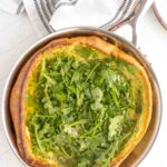 A baked Dutch baby pancake in a pan topped with fresh arugula, set on a white surface with a striped kitchen towel nearby.