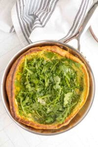 A baked Dutch baby pancake in a pan topped with fresh arugula, set on a white surface with a striped kitchen towel nearby.