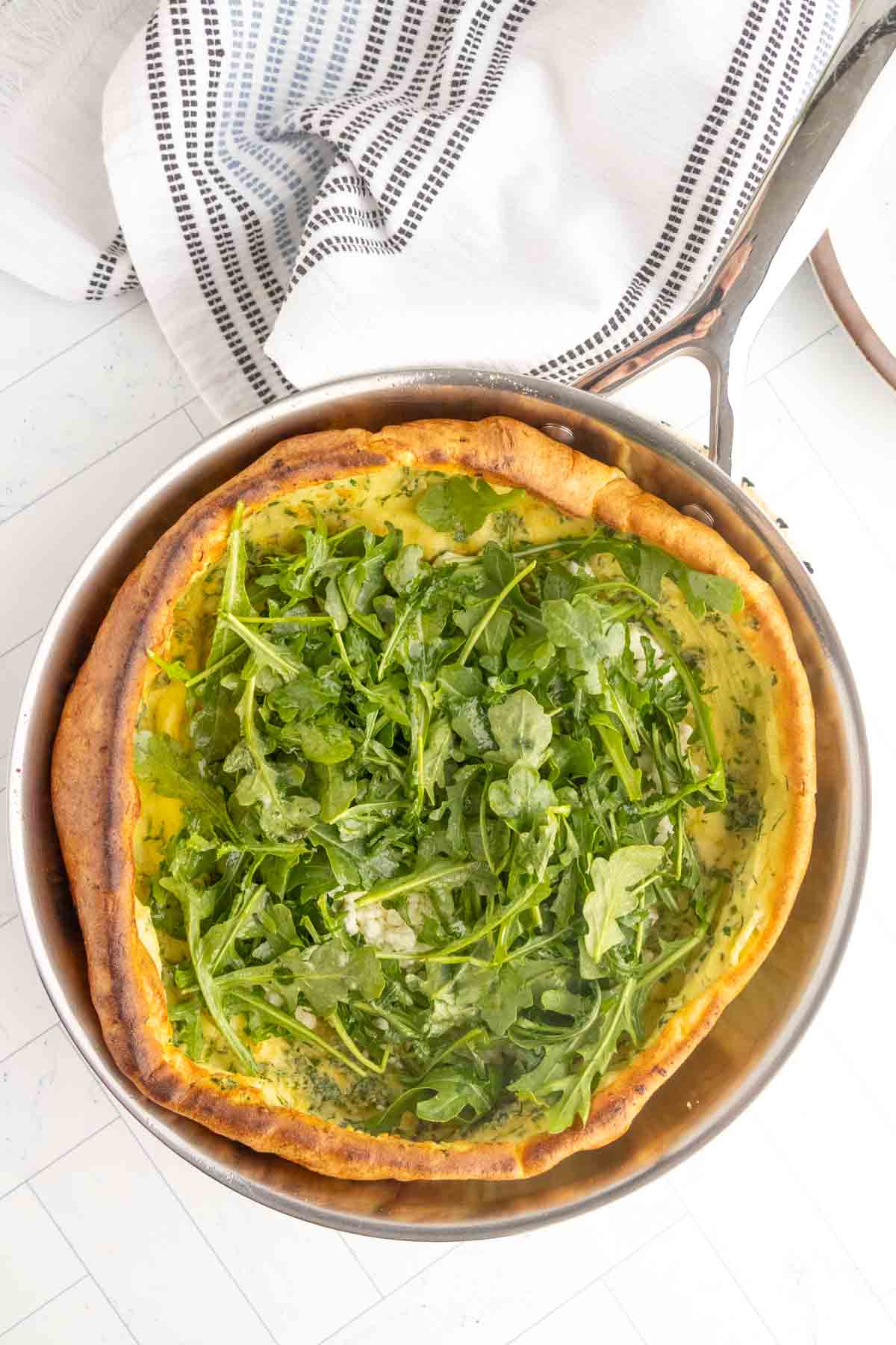 A baked Dutch baby pancake in a pan topped with fresh arugula, set on a white surface with a striped kitchen towel nearby.