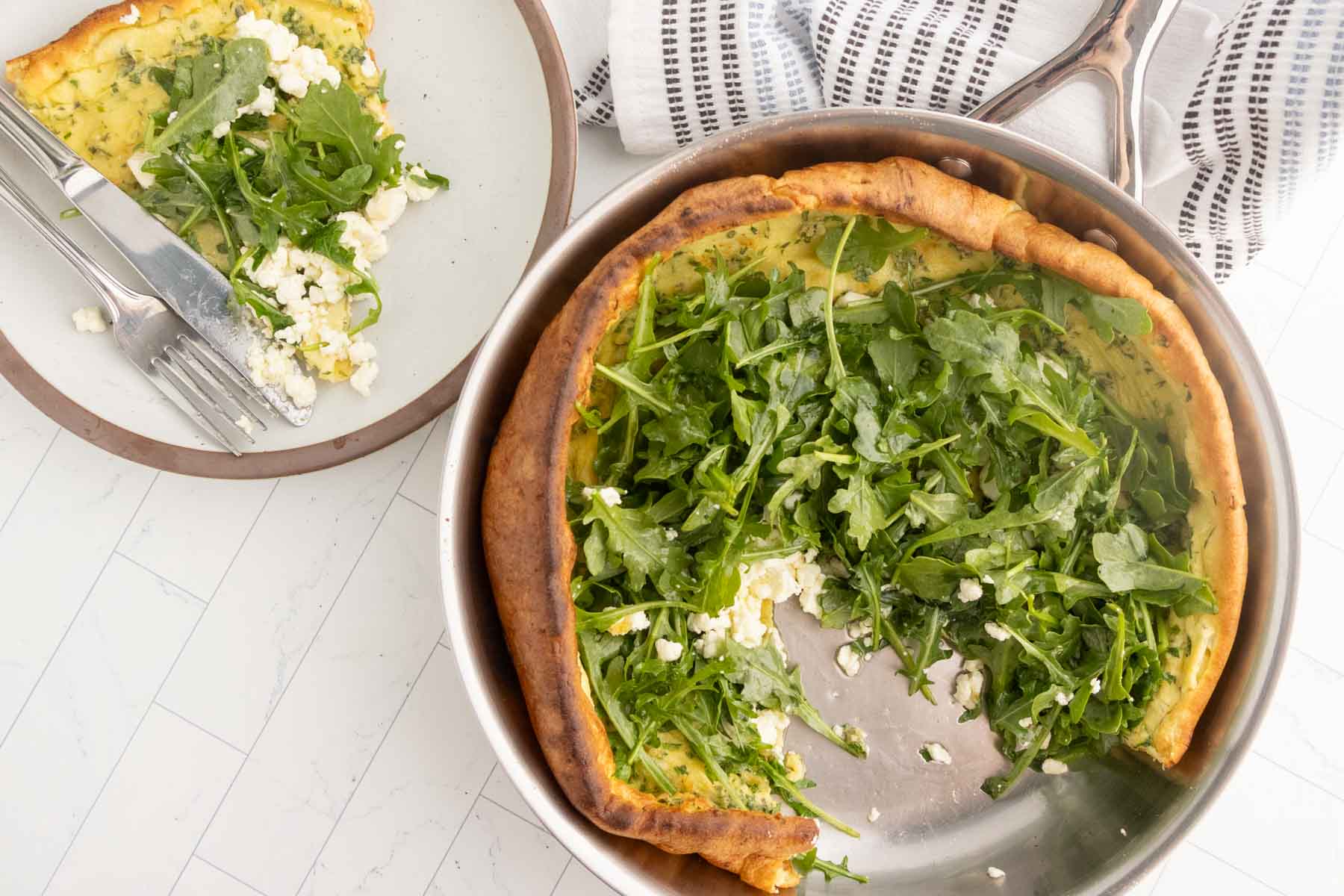 A baked dutch baby pancake in a pan topped with arugula and crumbled cheese, with a sliced piece served on a plate beside it.