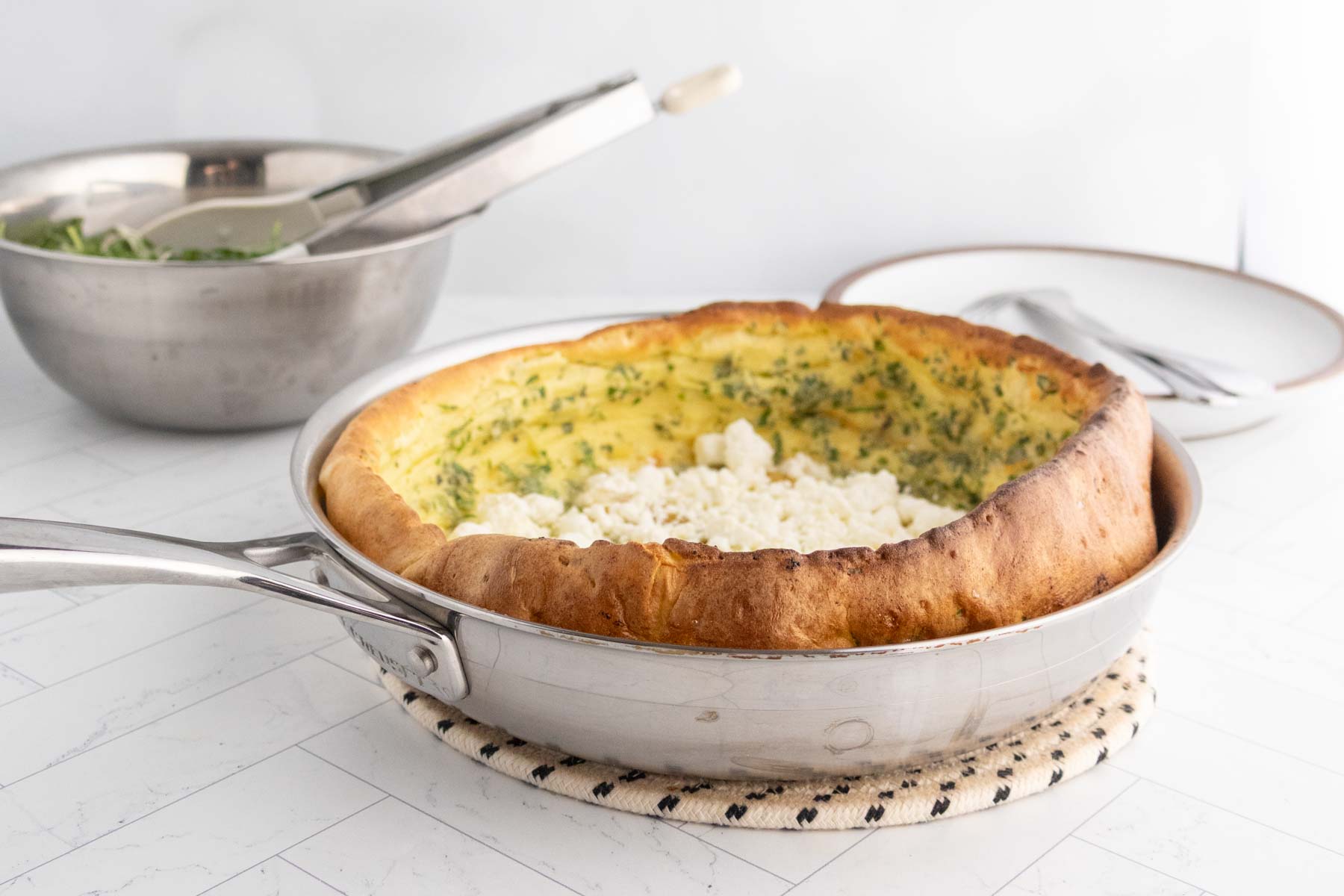 A baked savory Dutch baby pancake with herbs and crumbled cheese sits in a metal skillet on a trivet, with a mixing bowl and empty plates in the background.