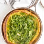 A savory Dutch baby pancake topped with fresh arugula sits in a metal skillet on a white surface, with a striped cloth partially visible.