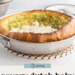 A baked Dutch baby pancake with chopped herbs and crumbled goat cheese sits in a metal pan on a trivet.