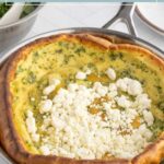 A baked savory Dutch baby pancake topped with crumbled cheese, served in a metal pan with greens visible in the background.