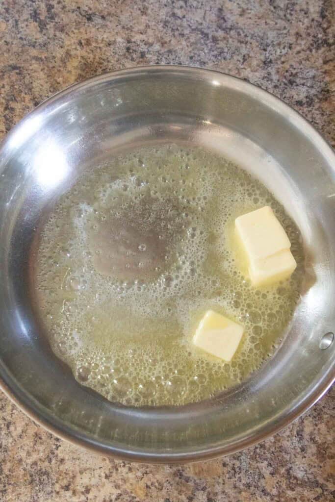 A stainless steel pan with melted butter and two unmelted butter cubes on a speckled countertop.