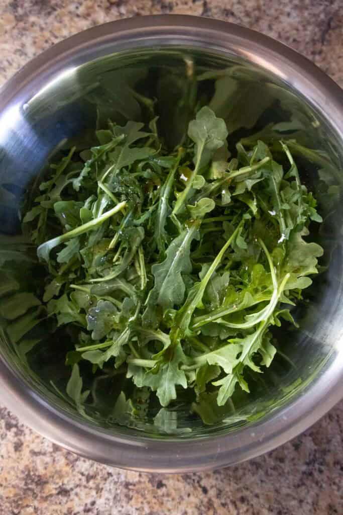 A stainless steel bowl filled with fresh arugula leaves sits on a speckled kitchen countertop.