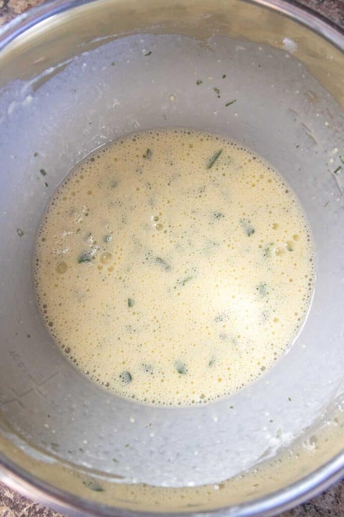A stainless steel bowl containing a frothy egg mixture with chopped green herbs.