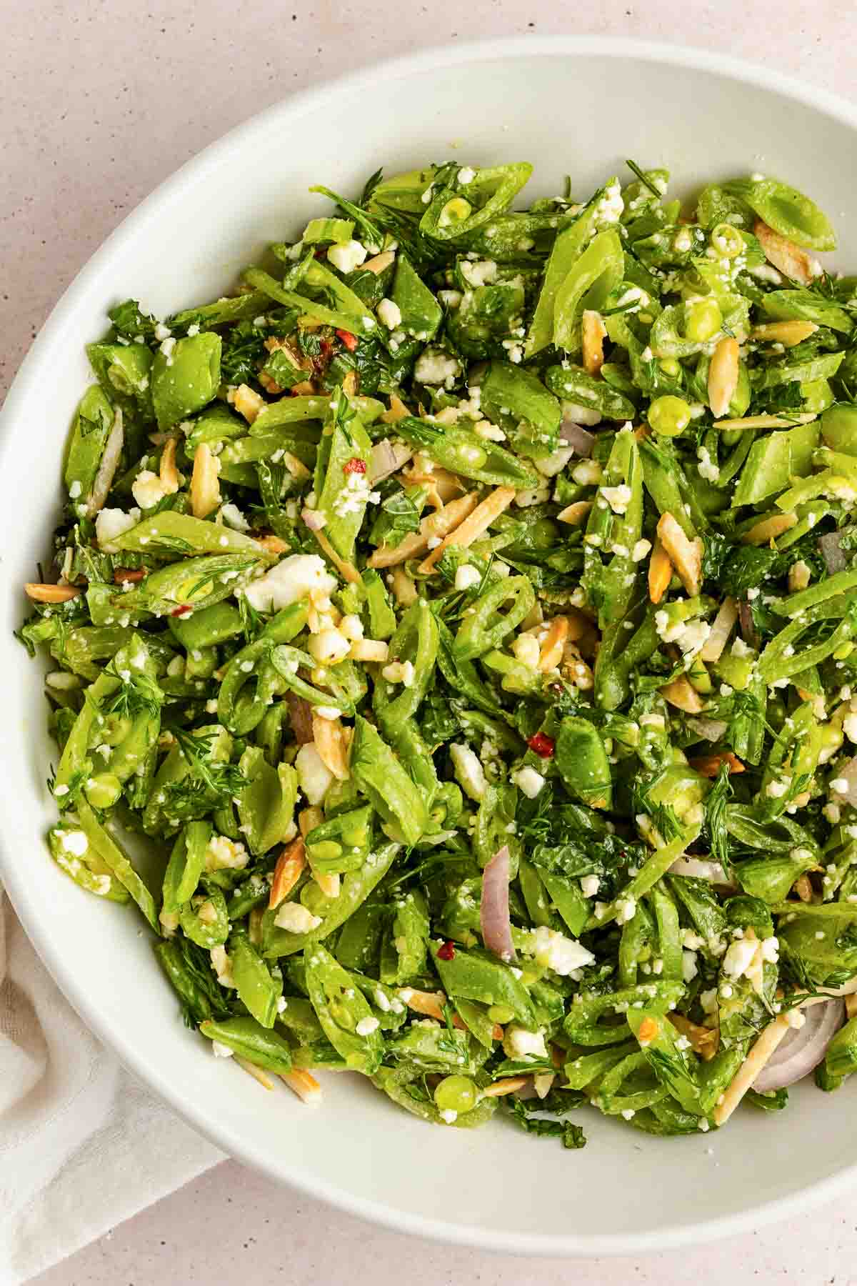 A bowl of salad with sliced snap peas, feta cheese, red onion, slivered almonds, and fresh herbs.