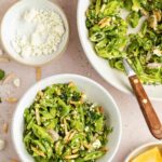 A bowl of sugar snap pea salad with feta cheese, slivered almonds, and herbs sits beside a larger serving dish, a small bowl of feta, and lemon wedges on a light surface.
