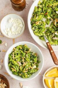 A bowl of sugar snap pea salad with feta cheese, slivered almonds, and herbs sits beside a larger serving dish, a small bowl of feta, and lemon wedges on a light surface.