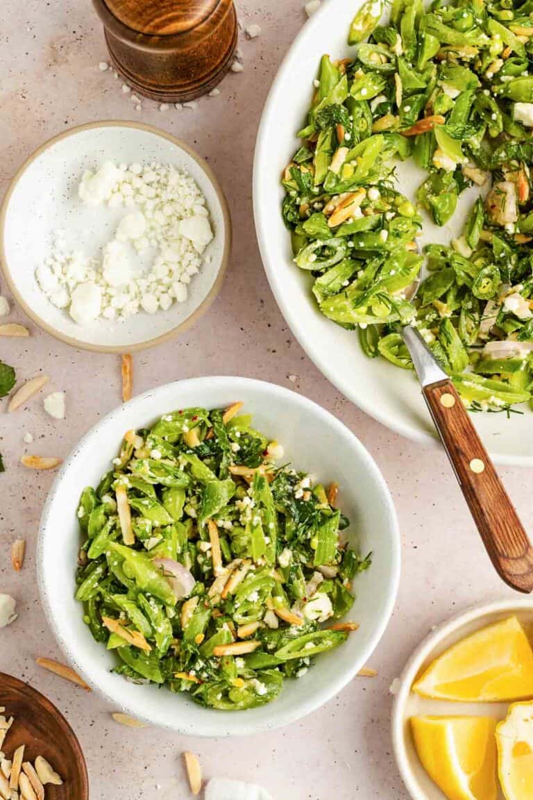 A bowl of sugar snap pea salad with feta cheese, slivered almonds, and herbs sits beside a larger serving dish, a small bowl of feta, and lemon wedges on a light surface.