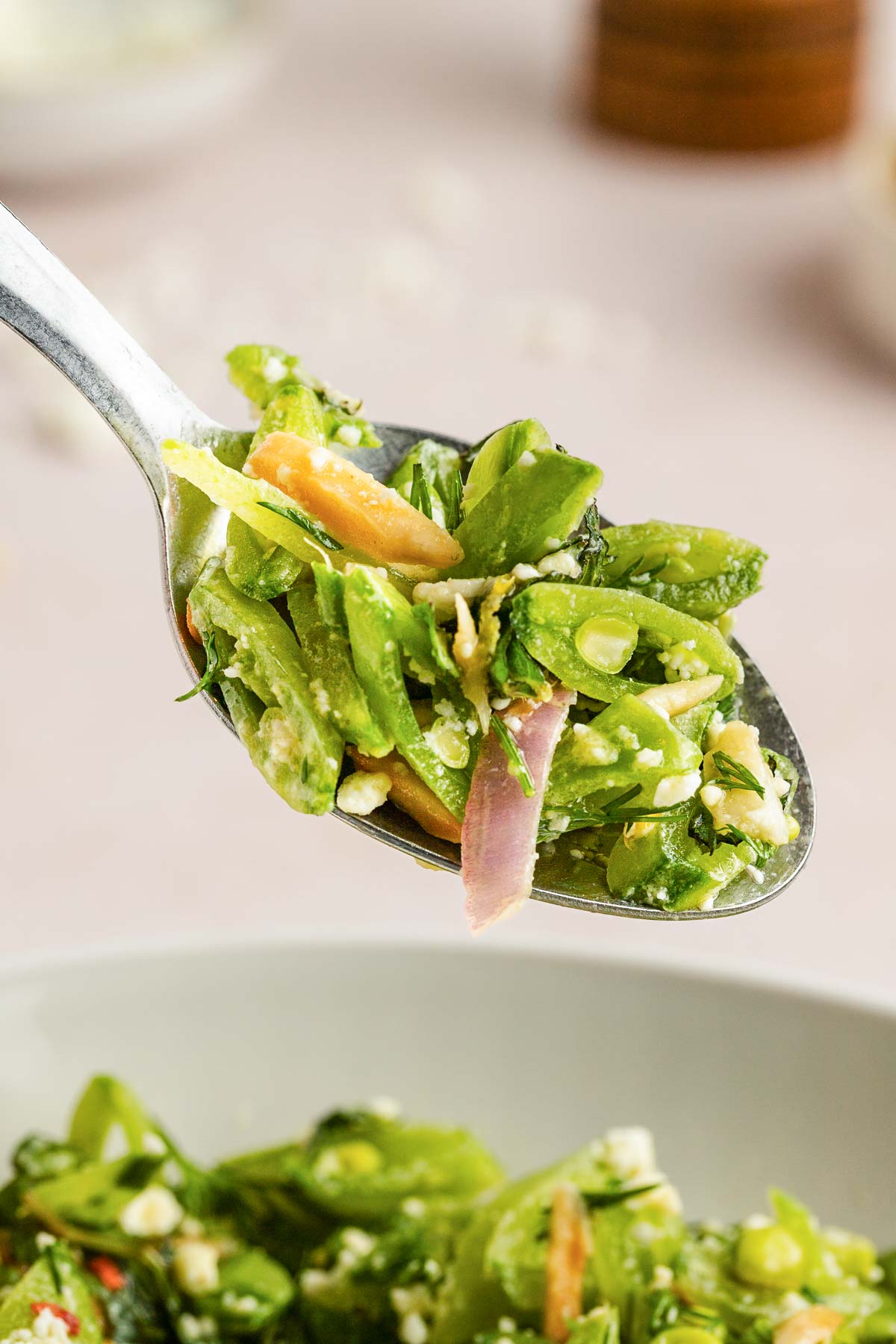 A close-up of a spoonful of green snap pea salad with sliced red onions, carrots, and crumbled cheese.