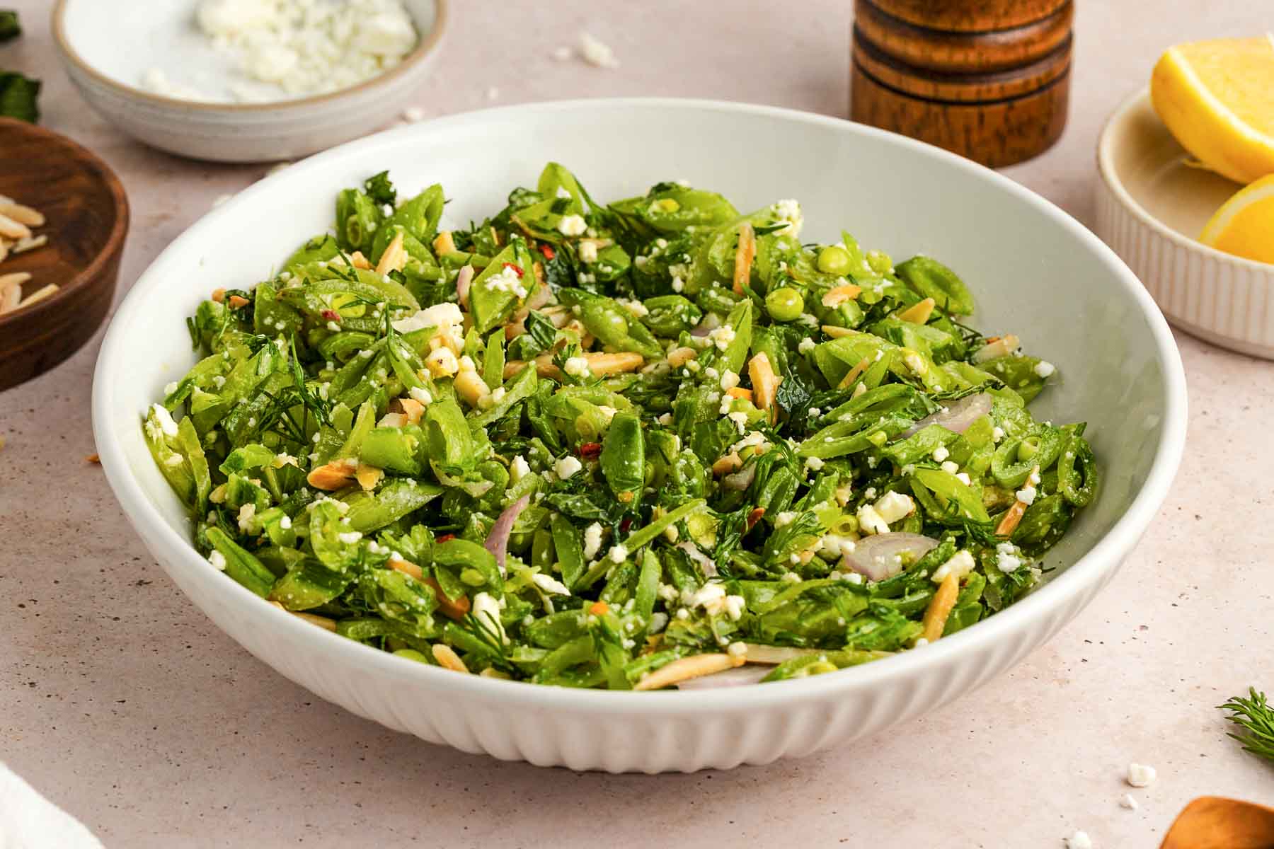 A white bowl filled with chopped green salad, sliced almonds, crumbled cheese, and herbs on a light-colored surface with lemon wedges and pepper grinder nearby.