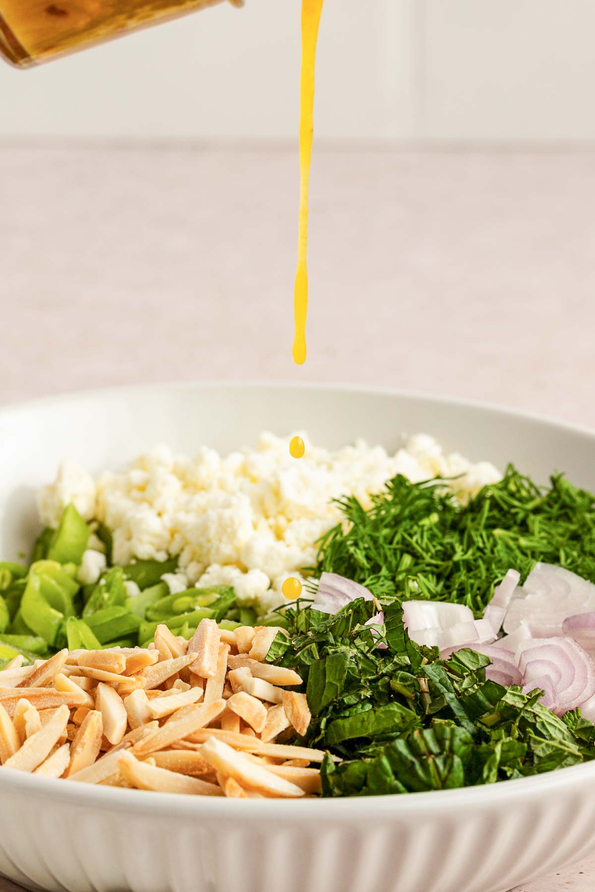 A close-up of a salad bowl with sliced almonds, chopped greens, onions, dill, feta cheese, and green beans, with dressing being drizzled from above.