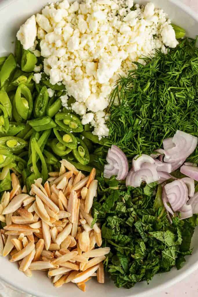 A bowl containing crumbled feta cheese, chopped dill, sliced shallots, sliced sugar snap peas, slivered almonds, and chopped fresh mint.