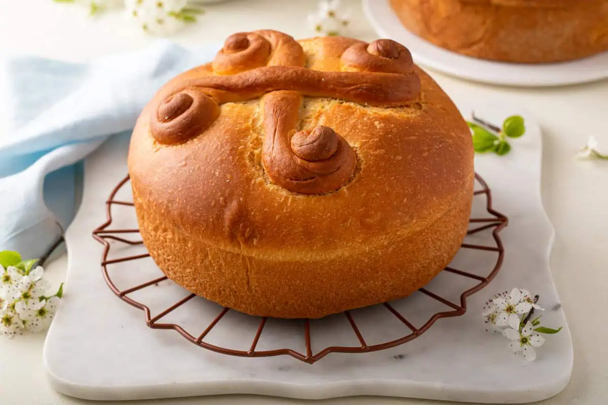 A round loaf of bread with decorative swirls on top, placed on a cooling rack over a marble surface, surrounded by small white flowers.