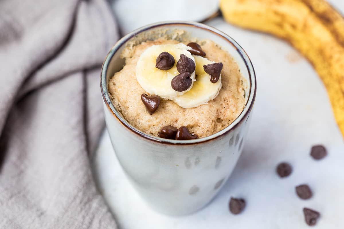 A ceramic mug filled with banana mug cake topped with banana slices and chocolate chips, placed on a light surface next to a napkin and a banana.