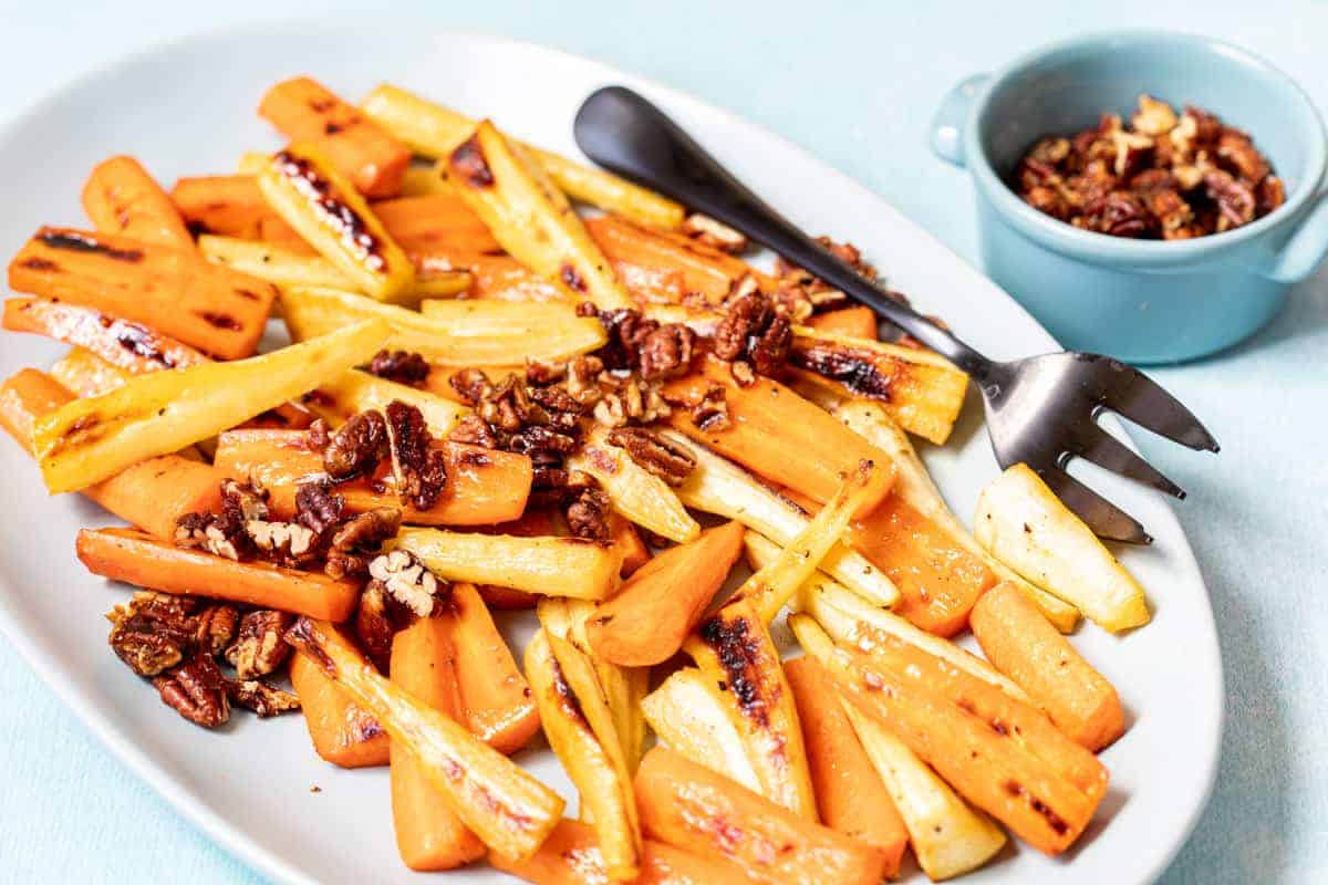 Roasted carrot and parsnip sticks topped with chopped pecans are served on a white plate with a black serving fork; extra pecans are in a small blue bowl.