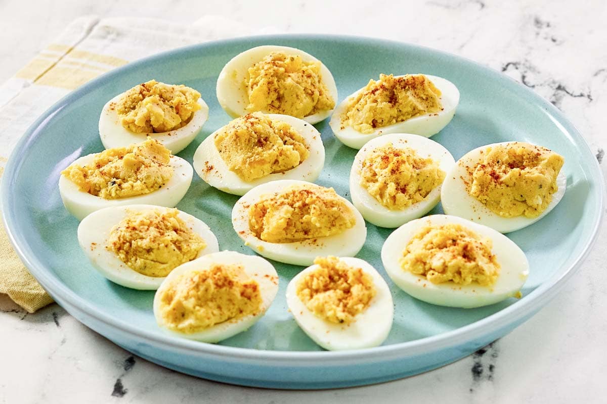 A blue plate with twelve deviled eggs, each topped with a sprinkle of paprika, sits on a marble surface next to a folded napkin.