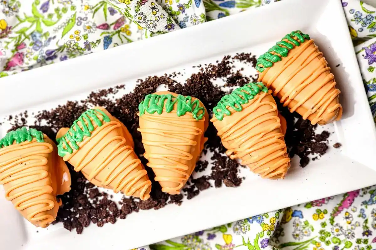 Five strawberries decorated to look like carrots with orange and green icing, arranged on crumbled chocolate cookies on a white rectangular plate.