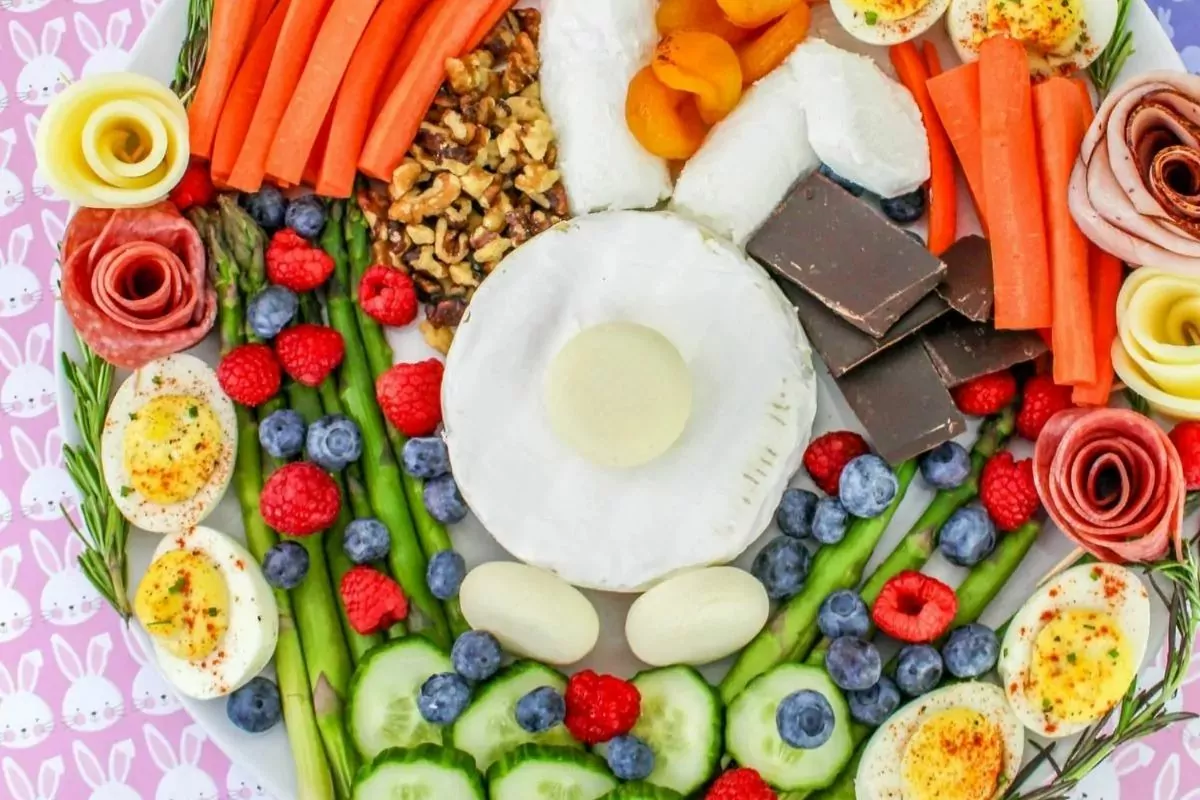 A platter arranged with sliced vegetables, boiled eggs, nuts, dried fruit, berries, chocolate, and cheese, styled to resemble a bunny face on a pink background with bunny prints.