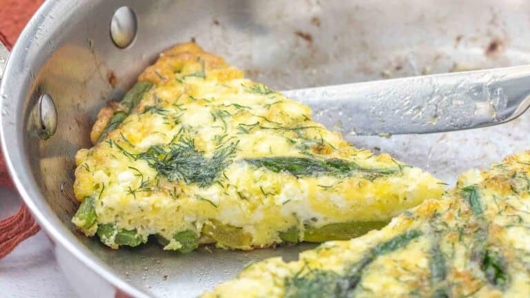 A slice of vegetable frittata with herbs is being served from a stainless steel pan with a knife.