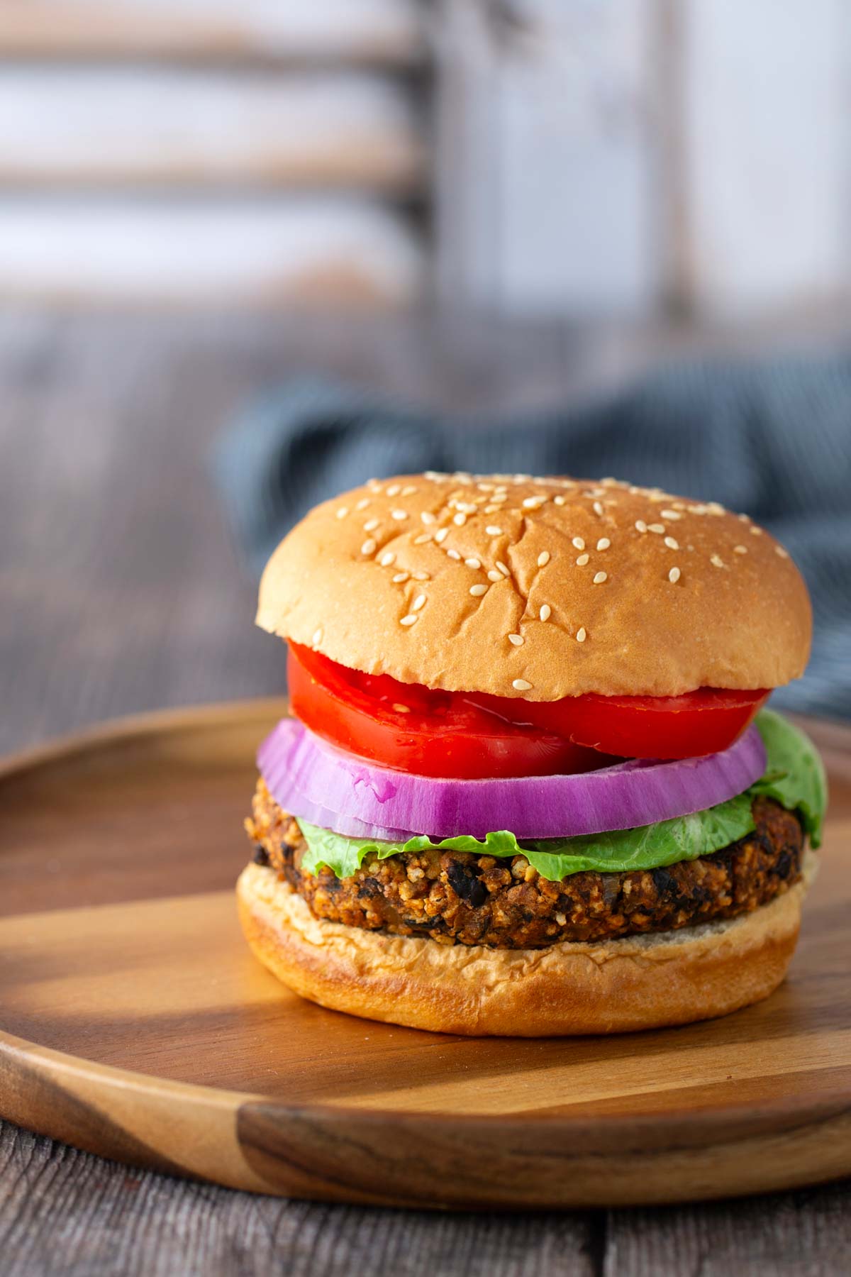 A veggie burger with lettuce, tomato, and red onion on a sesame seed bun sits on a wooden plate.