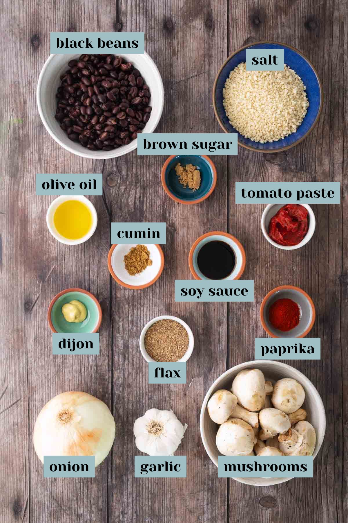 A variety of labeled ingredients on a wooden table, including black beans, salt, brown sugar, tomato paste, olive oil, cumin, soy sauce, paprika, dijon, flax, onion, garlic, and mushrooms.
