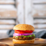 A veggie burger with lettuce, red onion, tomato, and a sesame seed bun on a wooden plate, with a blurred background.