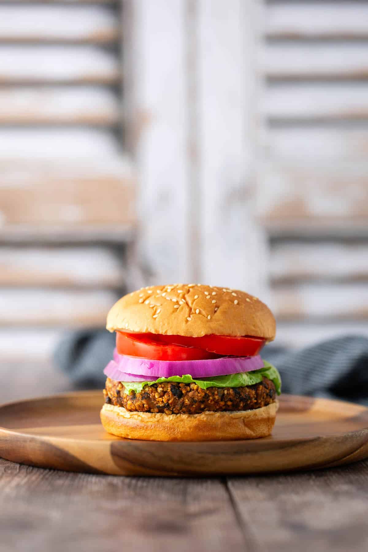 A veggie burger with lettuce, red onion, tomato, and a sesame seed bun on a wooden plate, with a blurred background.