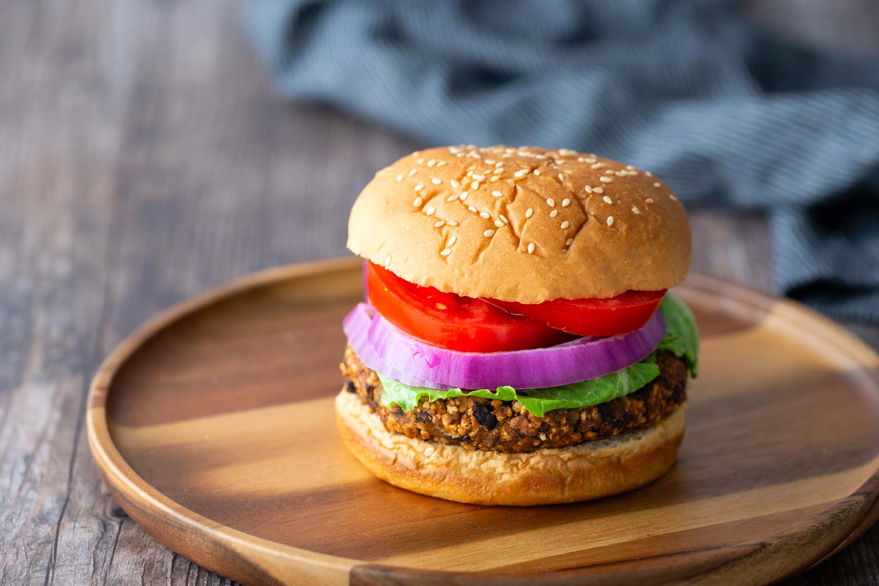 A veggie burger with lettuce, tomato, and red onion on a sesame seed bun sits on a wooden plate.