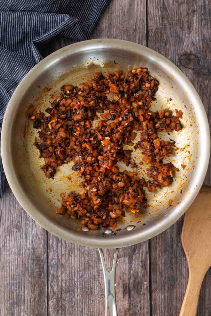 Chopped onions, carrots, and other vegetables sautéed in a stainless steel pan on a wooden surface with a wooden spatula beside it.