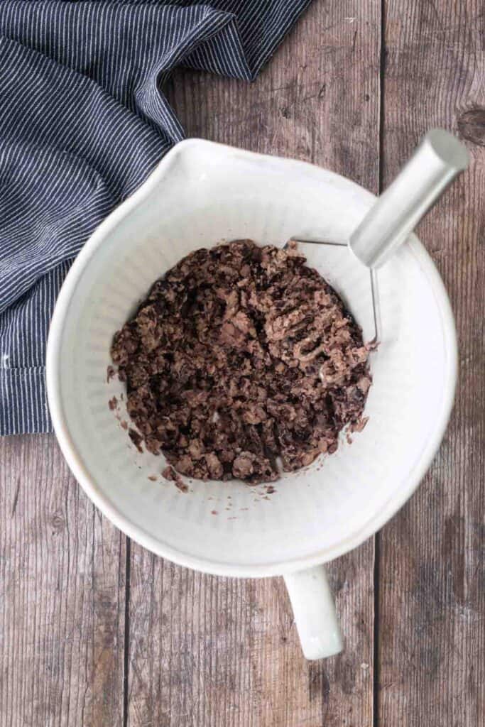 A white bowl containing mashed black beans with a metal potato masher, placed on a wooden surface next to a striped cloth.