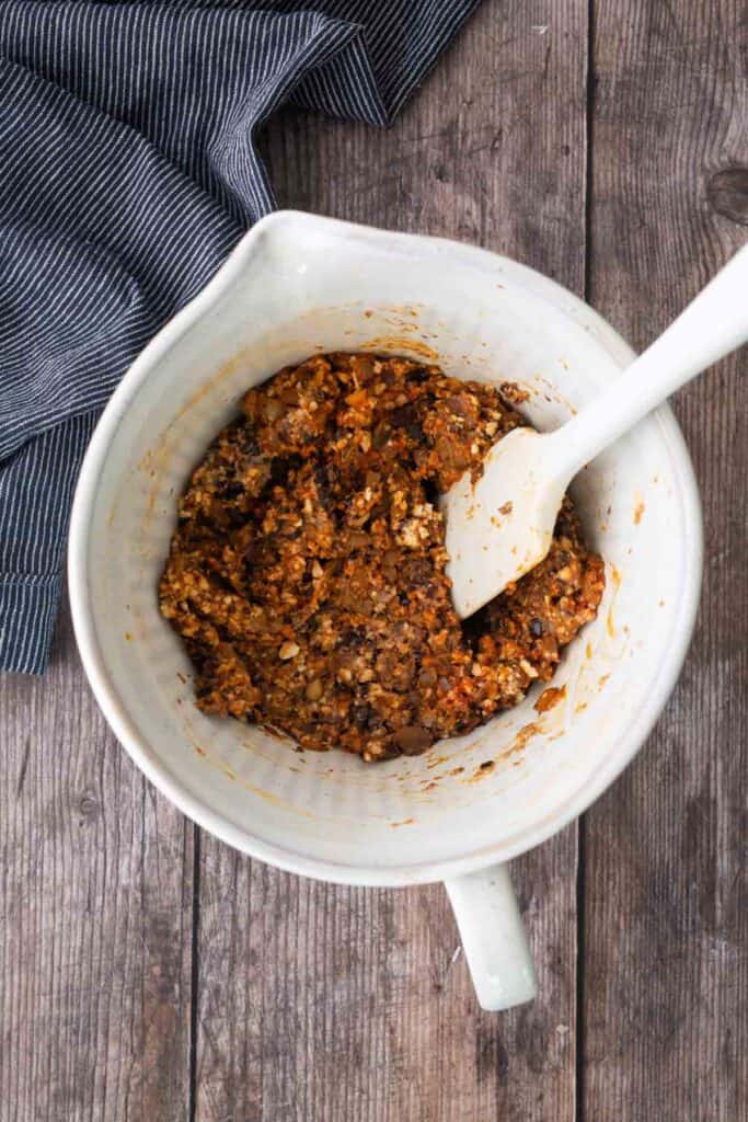 A large white mixing bowl contains a mixture of chopped vegetables and grains. A white spatula rests inside the bowl, and a striped cloth is nearby on a wooden surface.