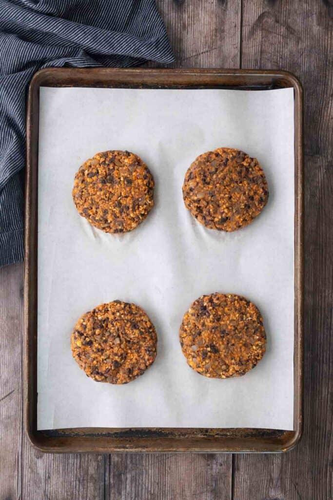 Four uncooked veggie burger patties on a parchment-lined baking sheet, placed on a wooden surface with a striped cloth in the upper left corner.