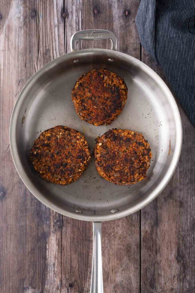 Three cooked veggie burger patties in a stainless steel pan on a wooden surface, with a dark striped cloth nearby.