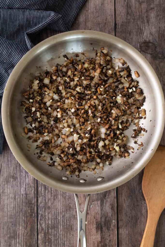 Chopped onions, garlic, and mushrooms sautéing in a stainless steel pan on a wooden surface, with a wooden spatula and a striped cloth nearby.