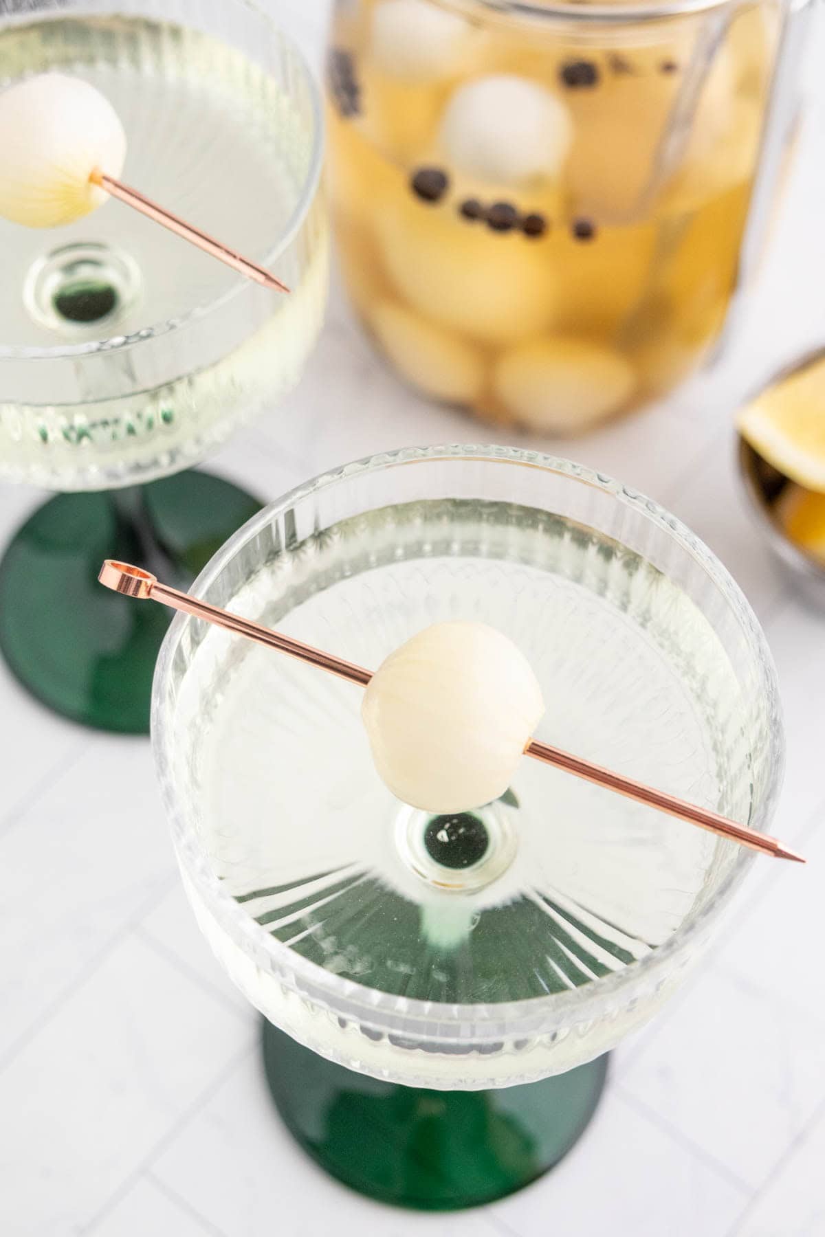 Two coupe glasses with clear cocktails, each garnished with a pickled onion on a cocktail pick, with a pitcher of similar liquid and onions in the background.