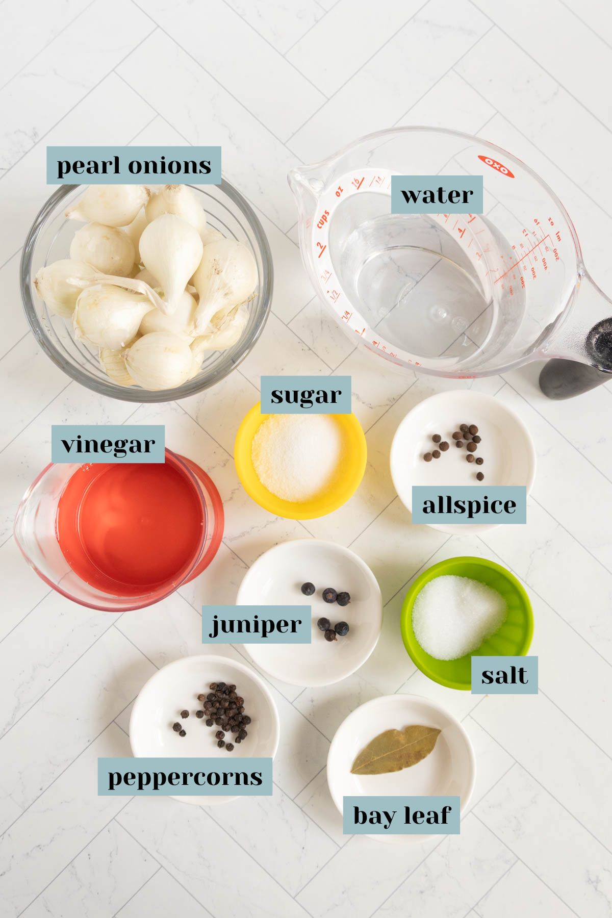 Bowls containing pearl onions, water, vinegar, sugar, allspice, juniper berries, salt, peppercorns, and a bay leaf are arranged on a white surface, each labeled.