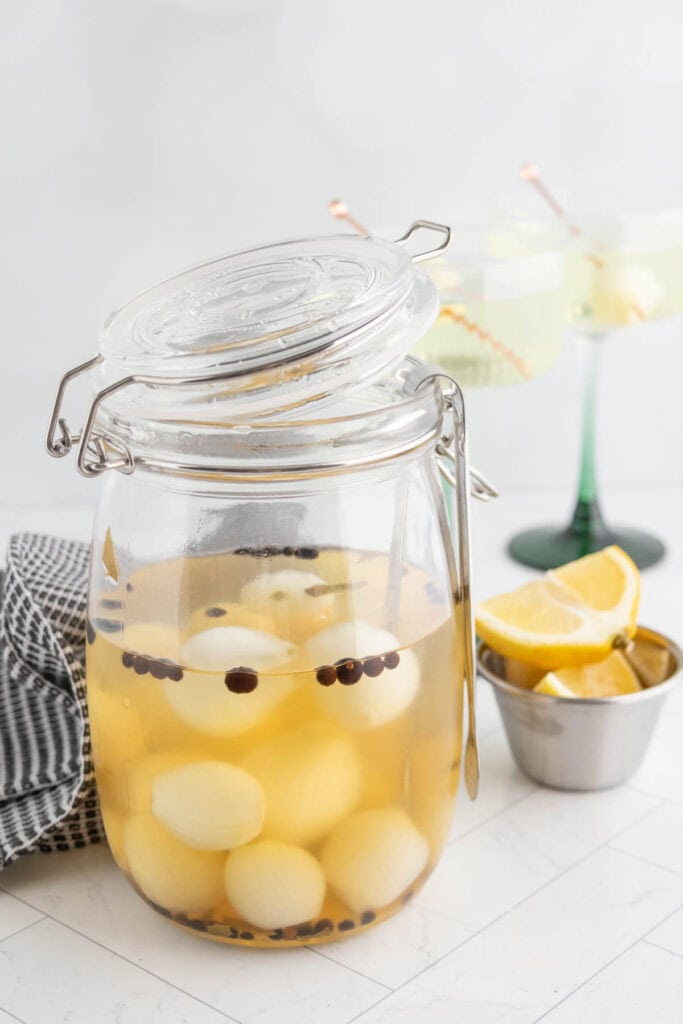 A glass jar filled with peeled pickled onions and spices sits on a counter next to lemon wedges and a striped cloth.