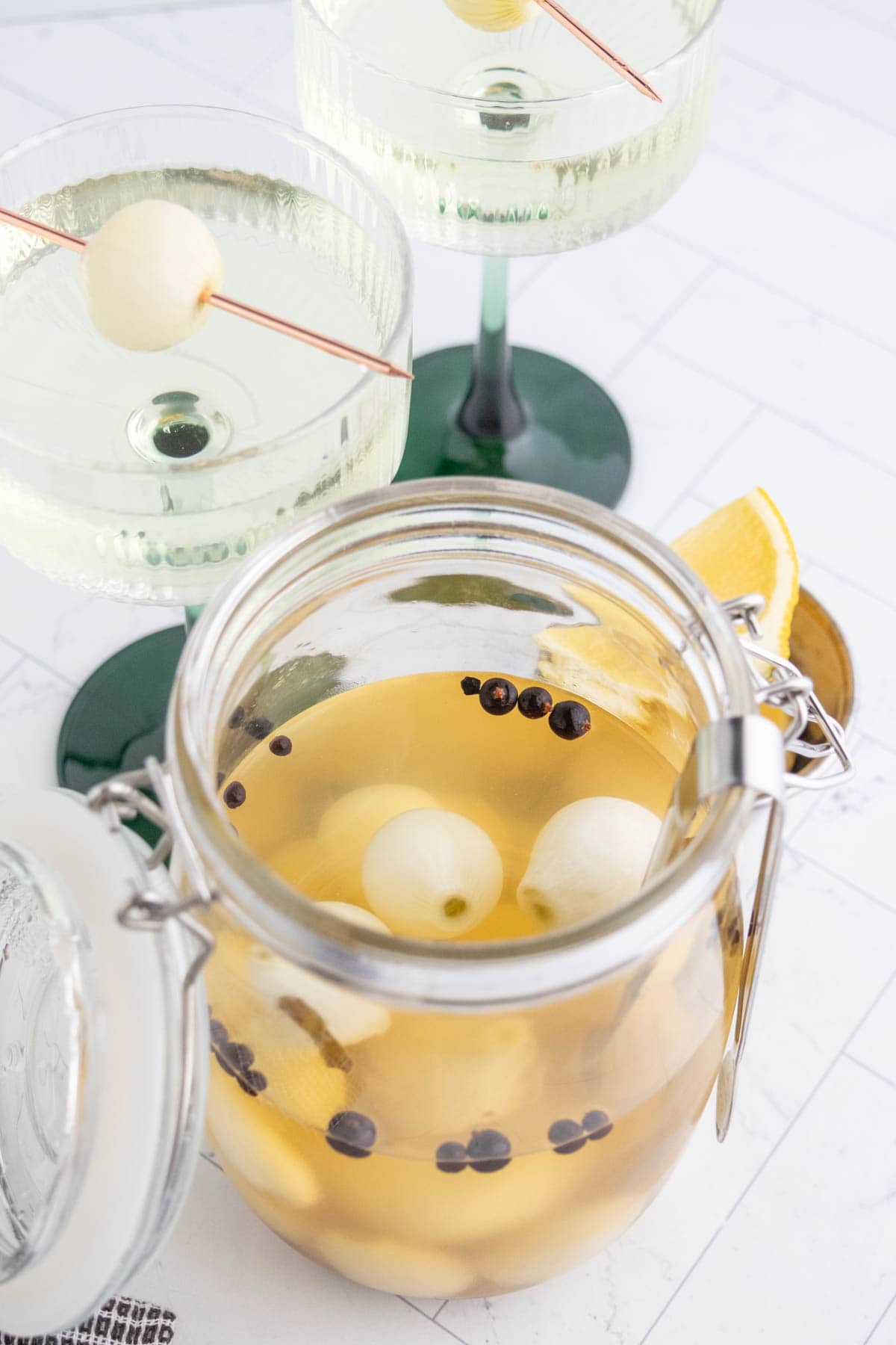 A jar of yellow liquid with melon balls, black peppercorns, and orange slices next to two glasses holding clear drinks garnished with melon balls on cocktail picks.